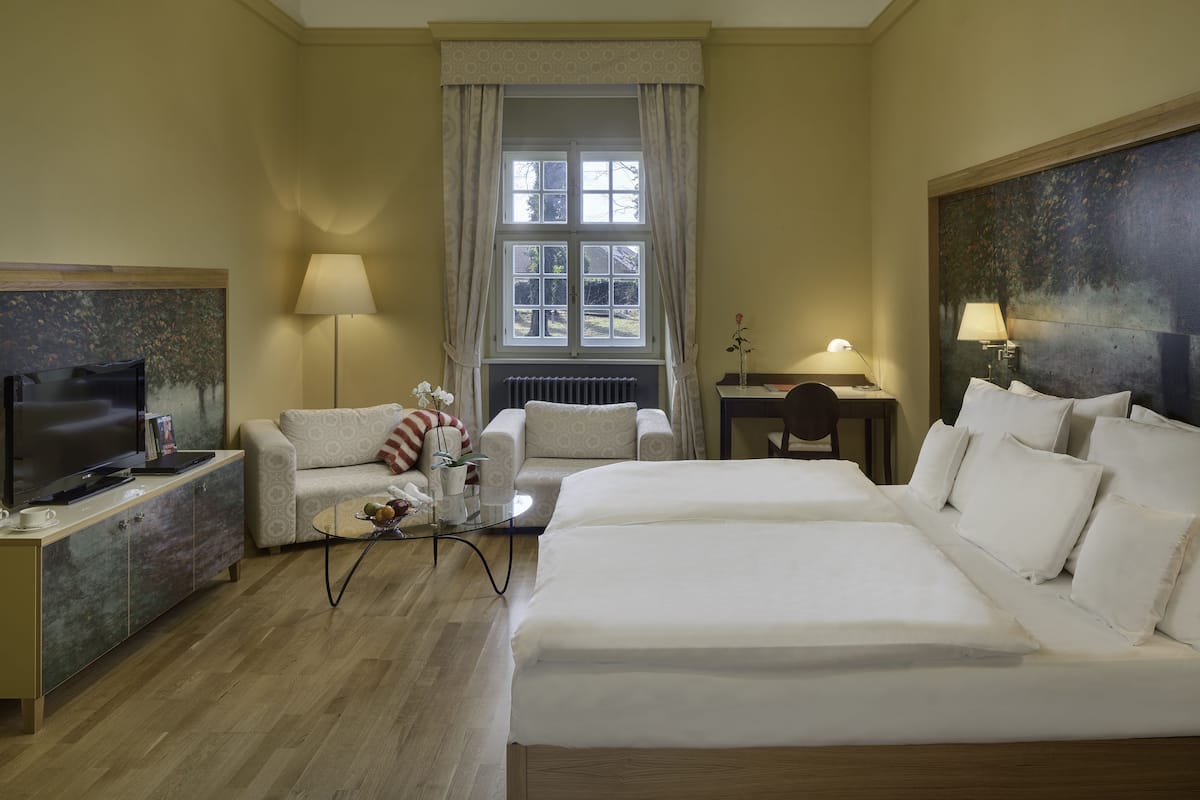 A hotel room with a large bed, two armchairs, a TV, and a desk with a lamp.