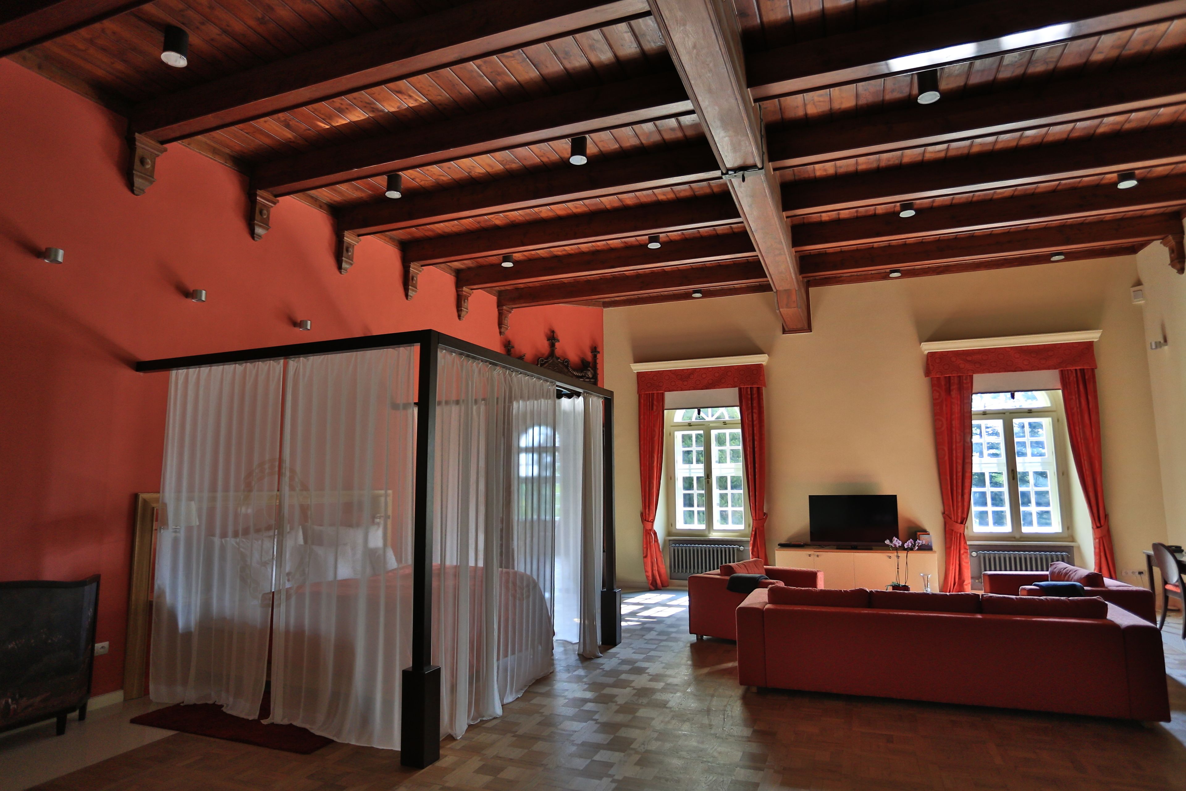 A room with a wooden ceiling, a bed with a canopy, a sofa, a television, and windows with curtains.