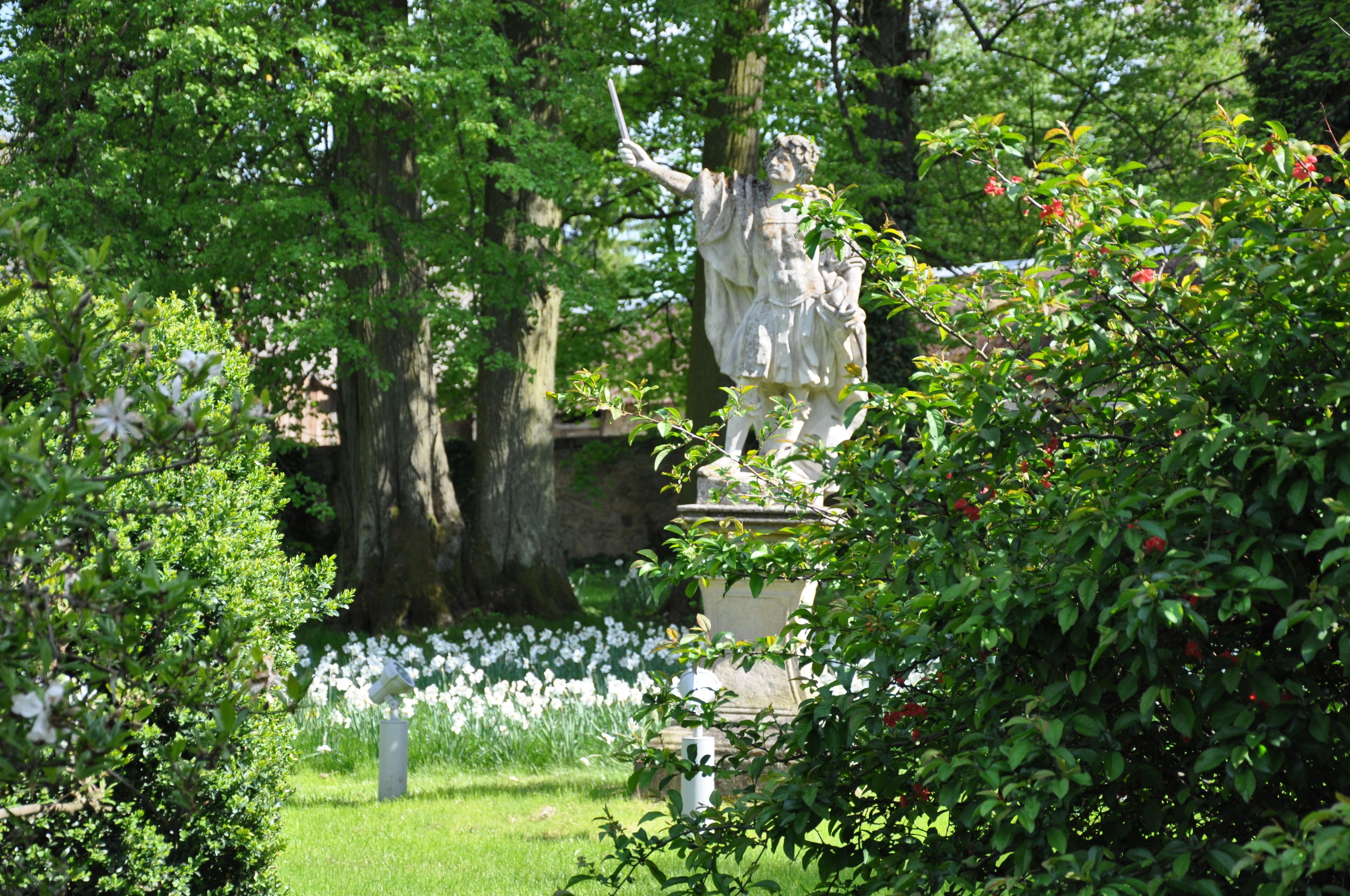 A statue in a lush garden with trees and white flowers.