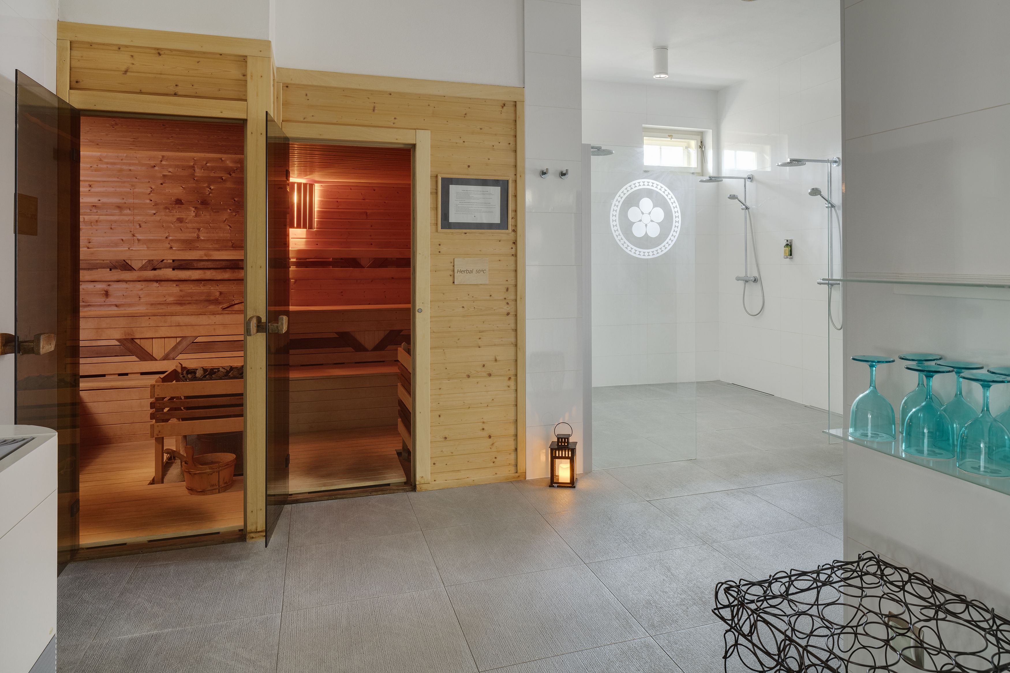 Una sala de sauna con bancos de madera y puerta de cristal, una zona de ducha con estantes de vidrio y un pequeño farol en el suelo.