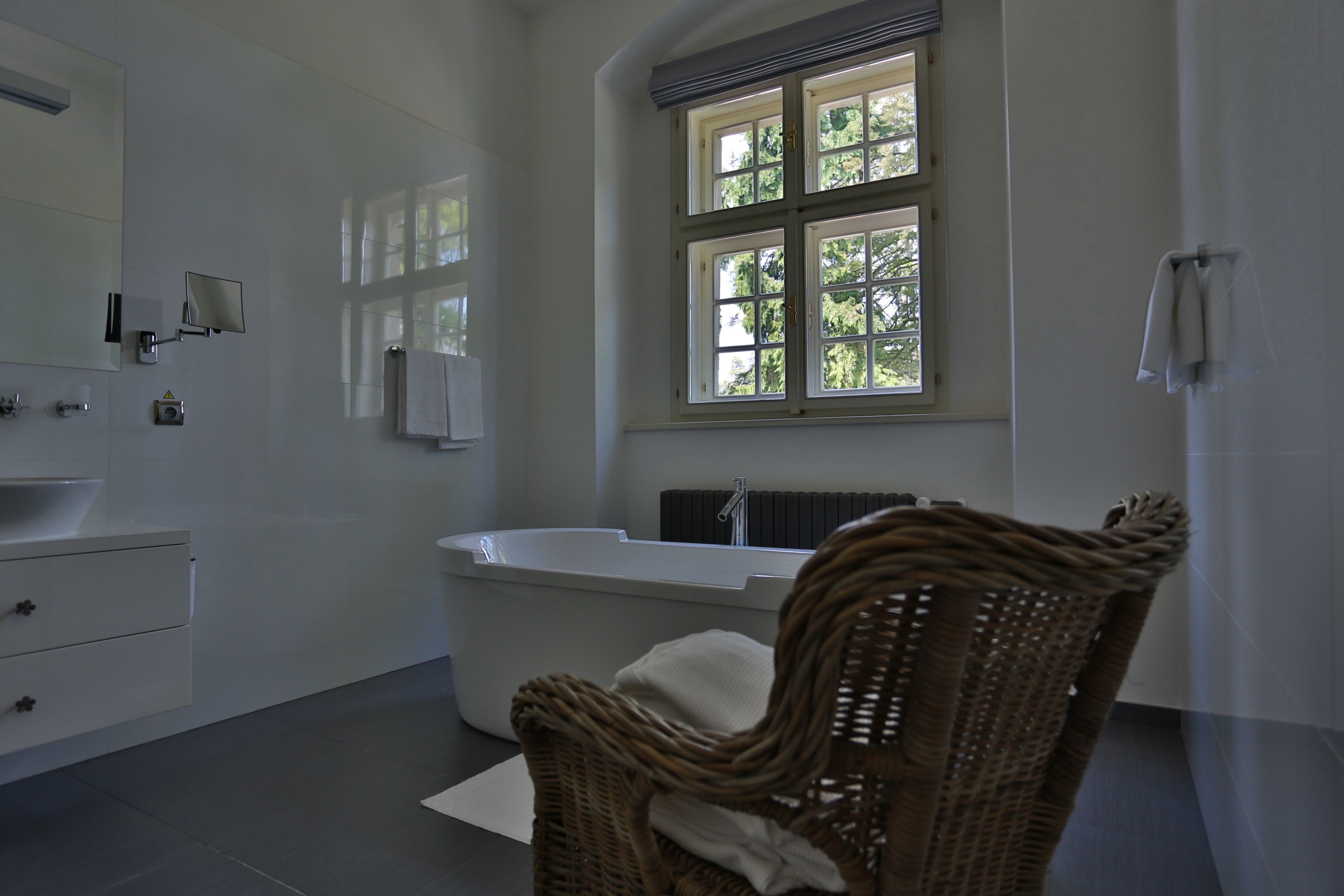 A modern bathroom with a large bathtub, a wicker chair, and a window with a view of trees.