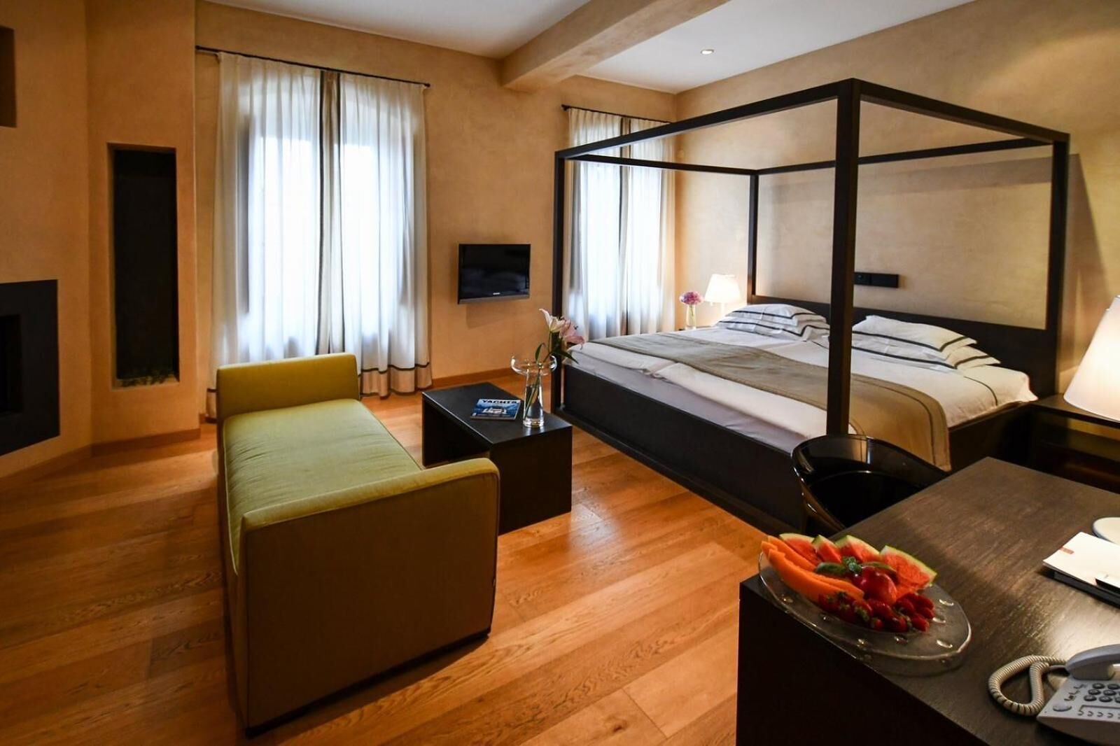 Et moderne hotelværelse med himmelseng, sofa, lille bord med vase og skål med frugt.