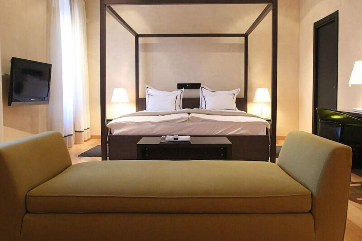 A hotel room with a four-poster bed, two bedside lamps, a flat-screen TV mounted on the wall, and a sofa.