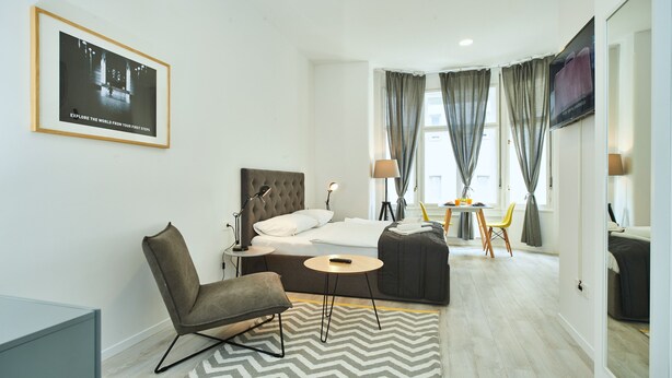 A modern hotel room with a bed, a chair, a desk, a TV, and a dining area with a table and chairs.