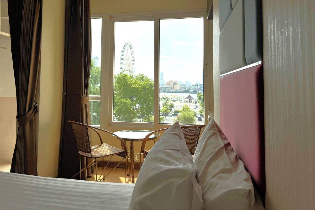 Hotellihuone, jossa on sänky, rottinkituoli, pieni pöytä ja ikkuna, josta avautuu näkymä huvipuiston pyörälaitteeseen ja kaupunkimaisemaan.