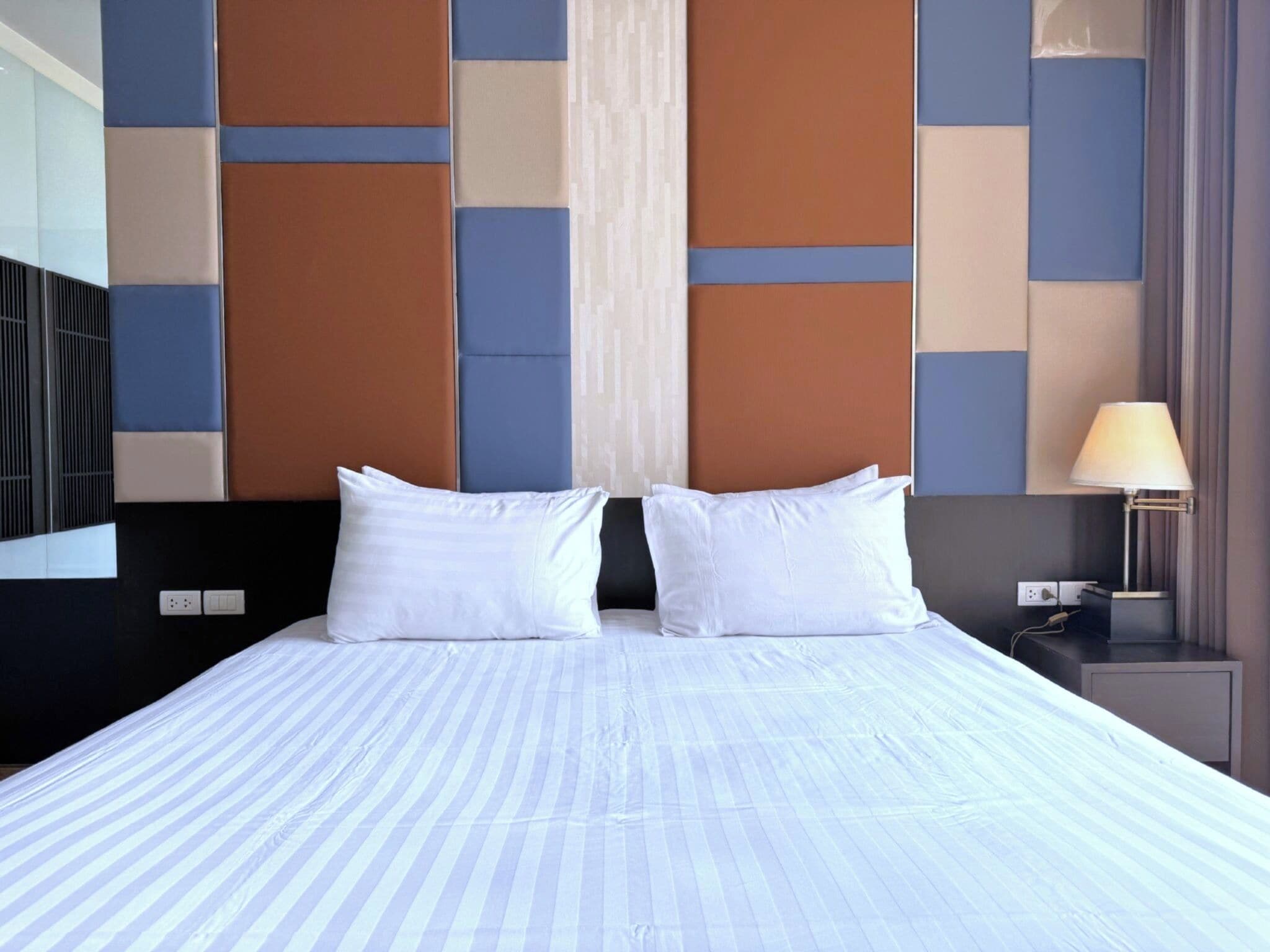 A hotel room with a large bed, two white pillows, a nightstand with a lamp, and a modern wall design.