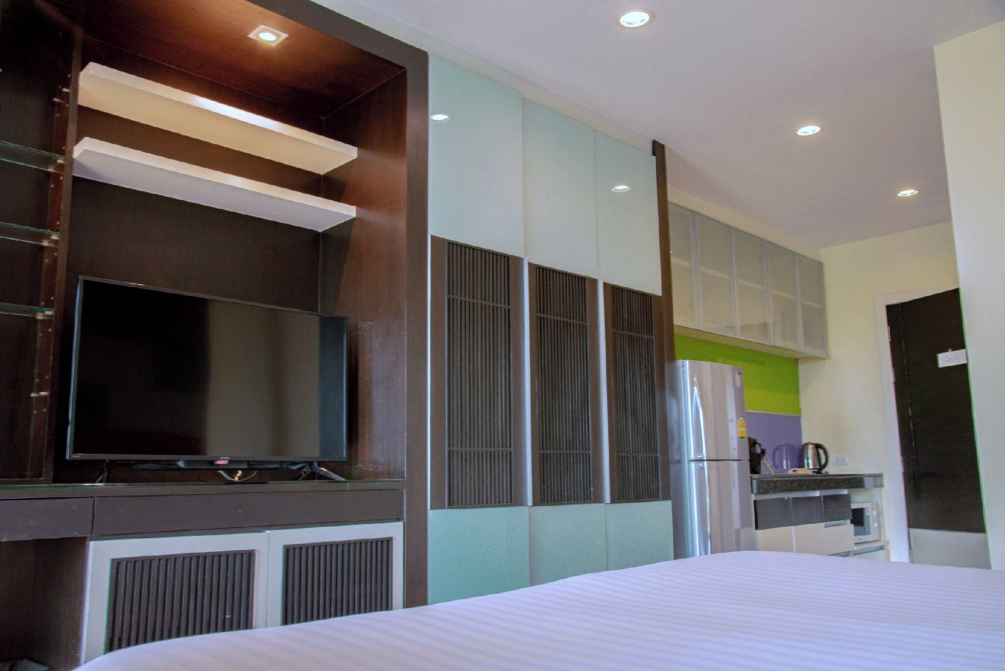 A modern hotel room with a large bed, a flat-screen TV mounted on a wooden cabinet, a refrigerator, and a kitchenette area.