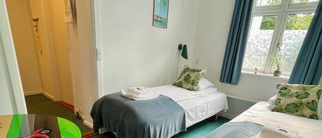 Una habitación con dos camas, un escritorio, una silla, un ventanal con cortinas y una puerta que da a otra habitación.