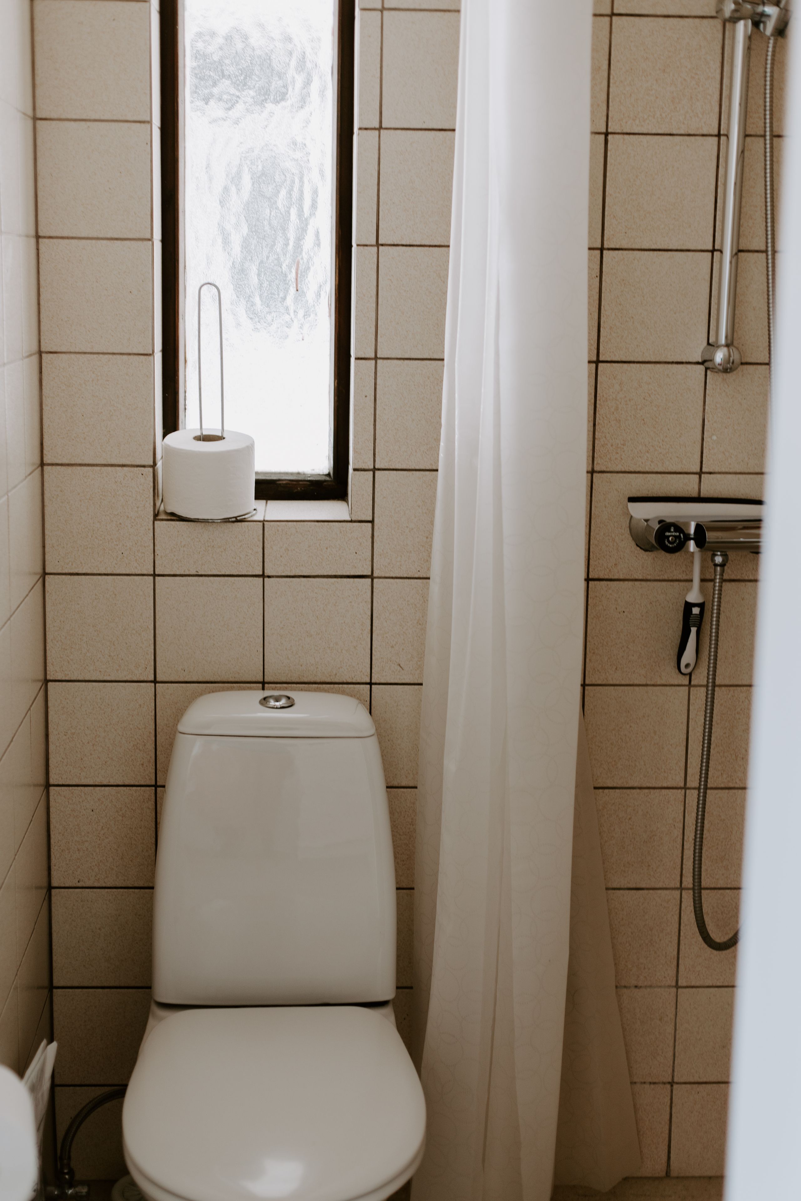A small bathroom with a toilet, a window with a paper towel dispenser, and a shower with a handheld showerhead.
