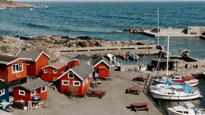 A coastal village with red wooden houses, a dock with boats, and a rocky shoreline. - Hotel Sandvig Havn (Allinge)