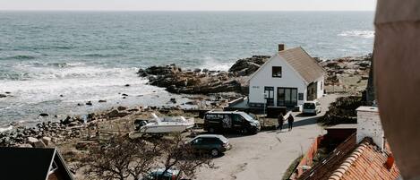 A coastal house with a view of the sea, a paved driveway, and parked cars.