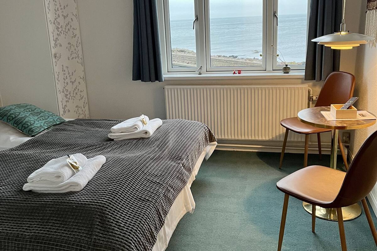 A hotel room with a bed, a chair, a table, and a view of the sea through a window.