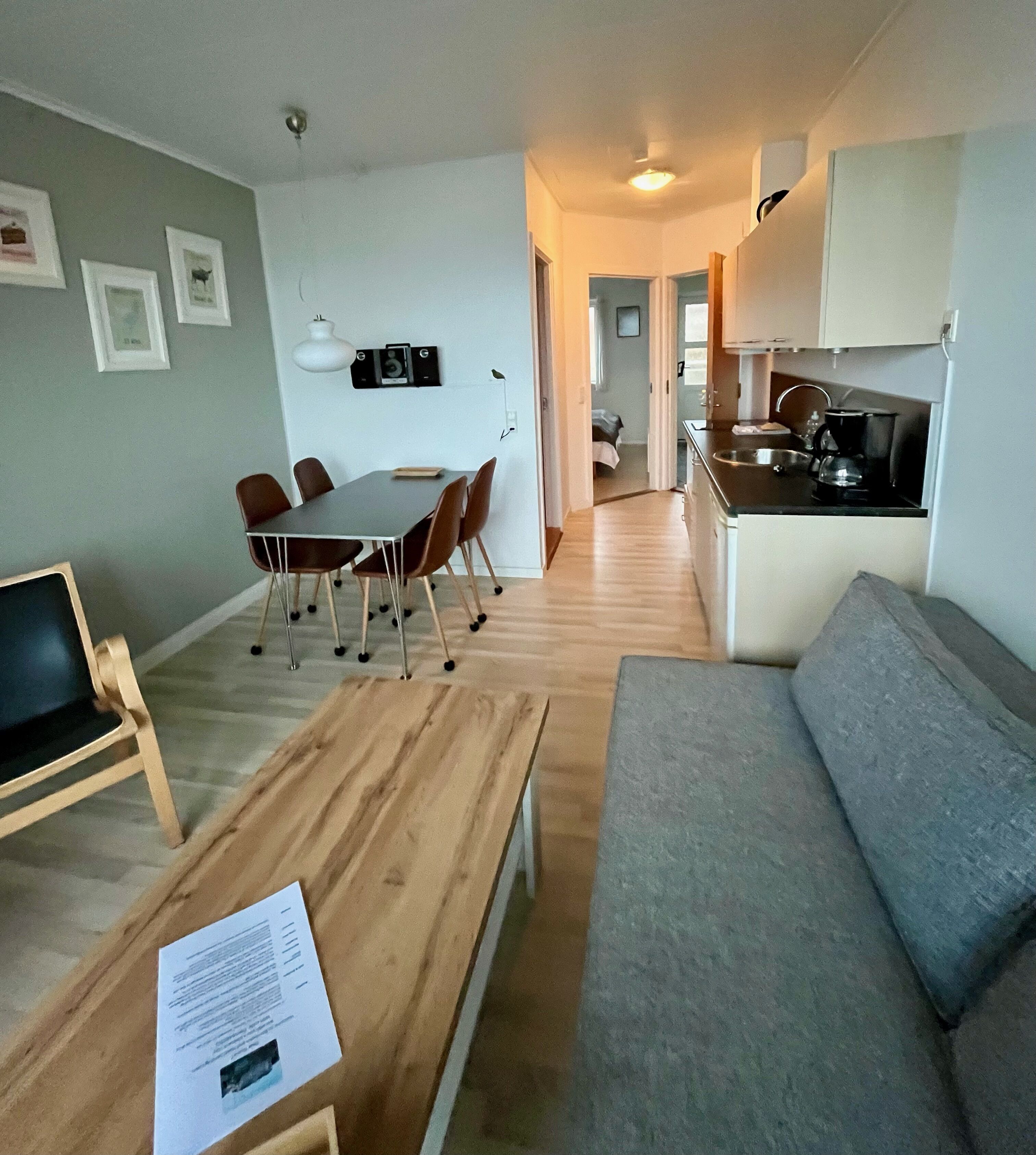 A modern living room with a grey sofa, a wooden coffee table, and a dining area with a table and chairs. A kitchen is visible in the background.