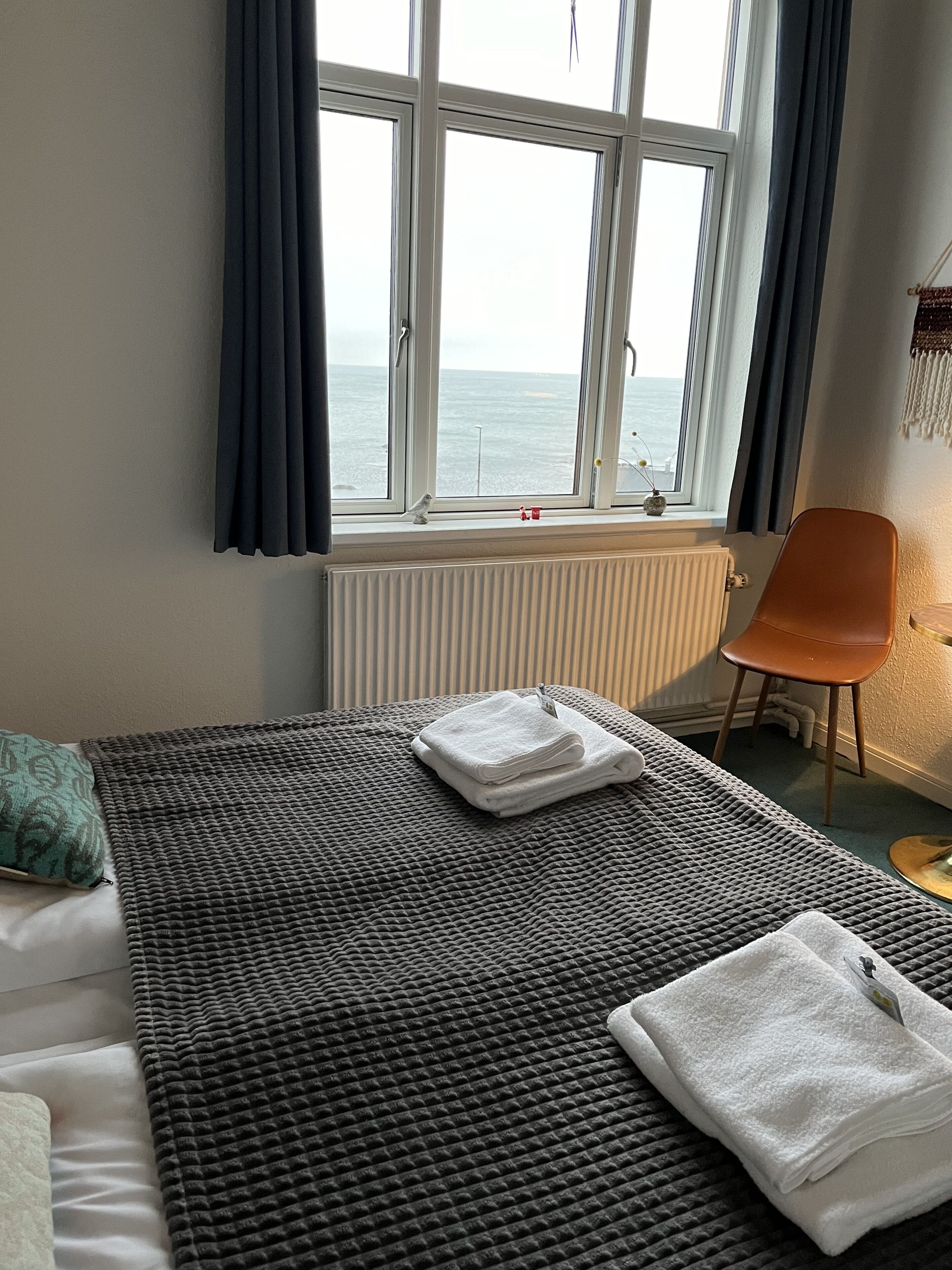 Habitación de hotel con cama, vista al mar a través de una ventana, una silla y un radiador.