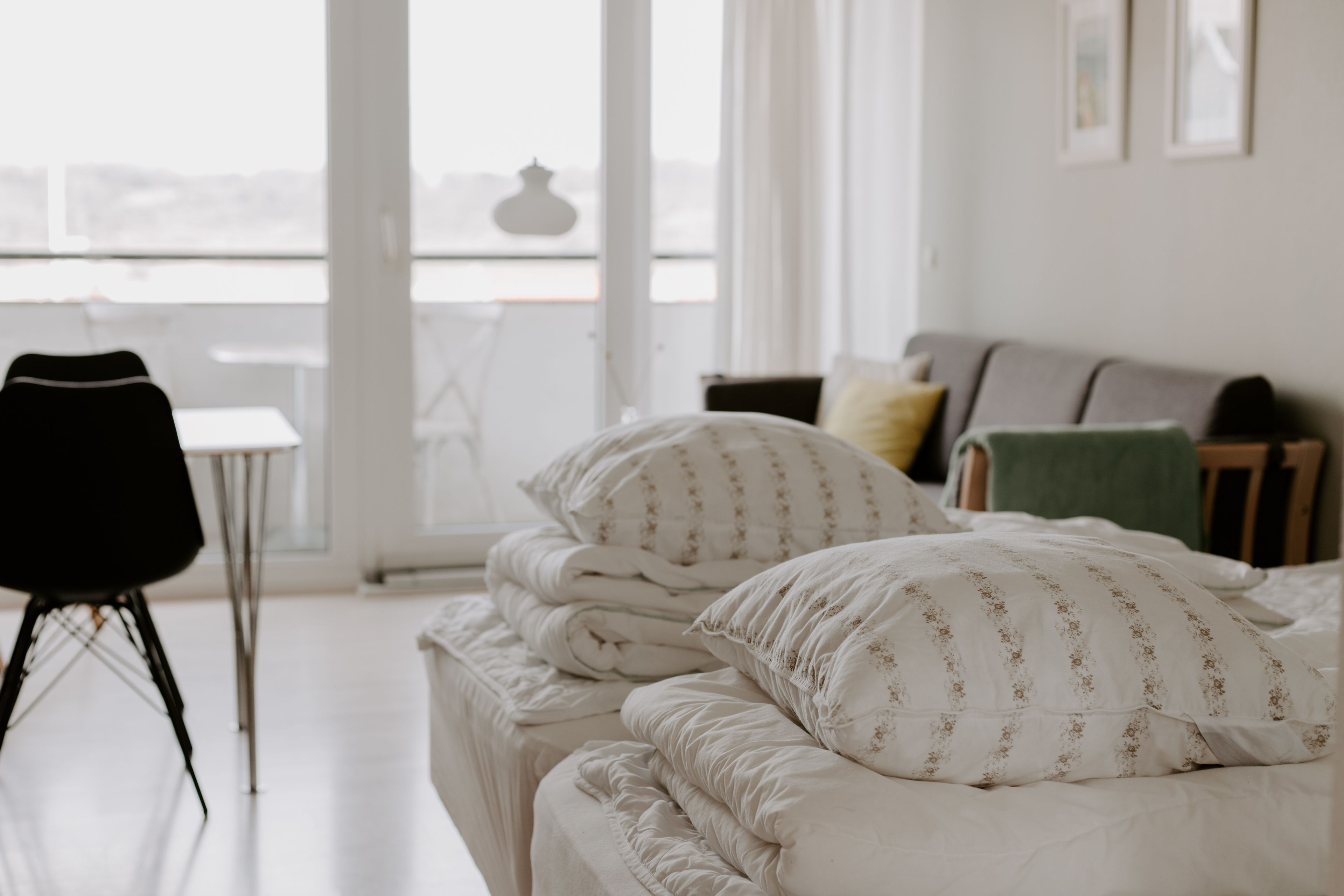 Un lit bien fait, avec du linge de lit blanc et des motifs floraux, une chaise noire et une petite table dans une pièce lumineuse offrant une vue dégagée grâce à de grandes fenêtres.
