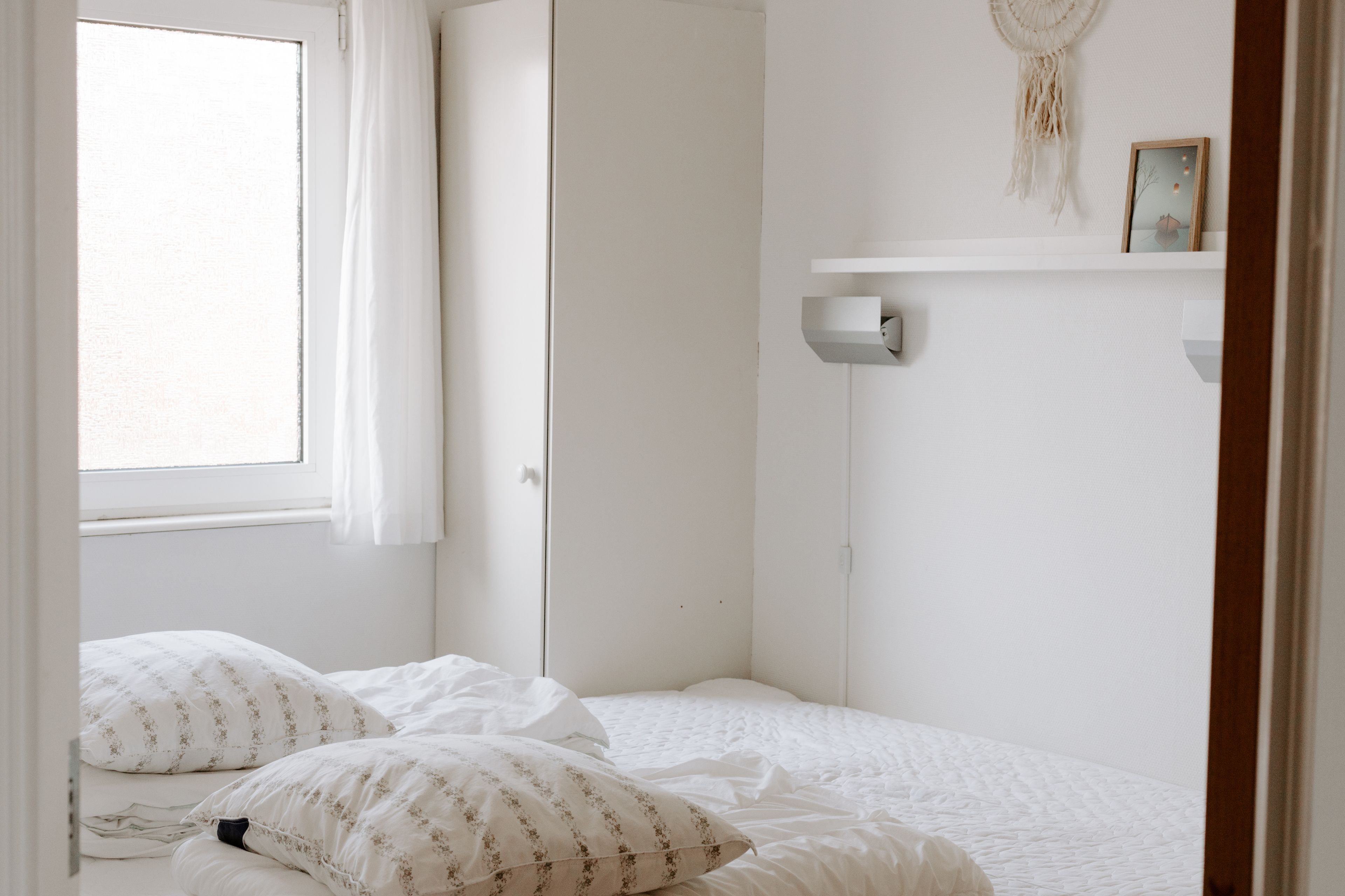 Une chambre avec un lit simple, du linge de lit blanc, une table de chevet, une étagère murale avec un tableau encadré et une fenêtre avec des rideaux.