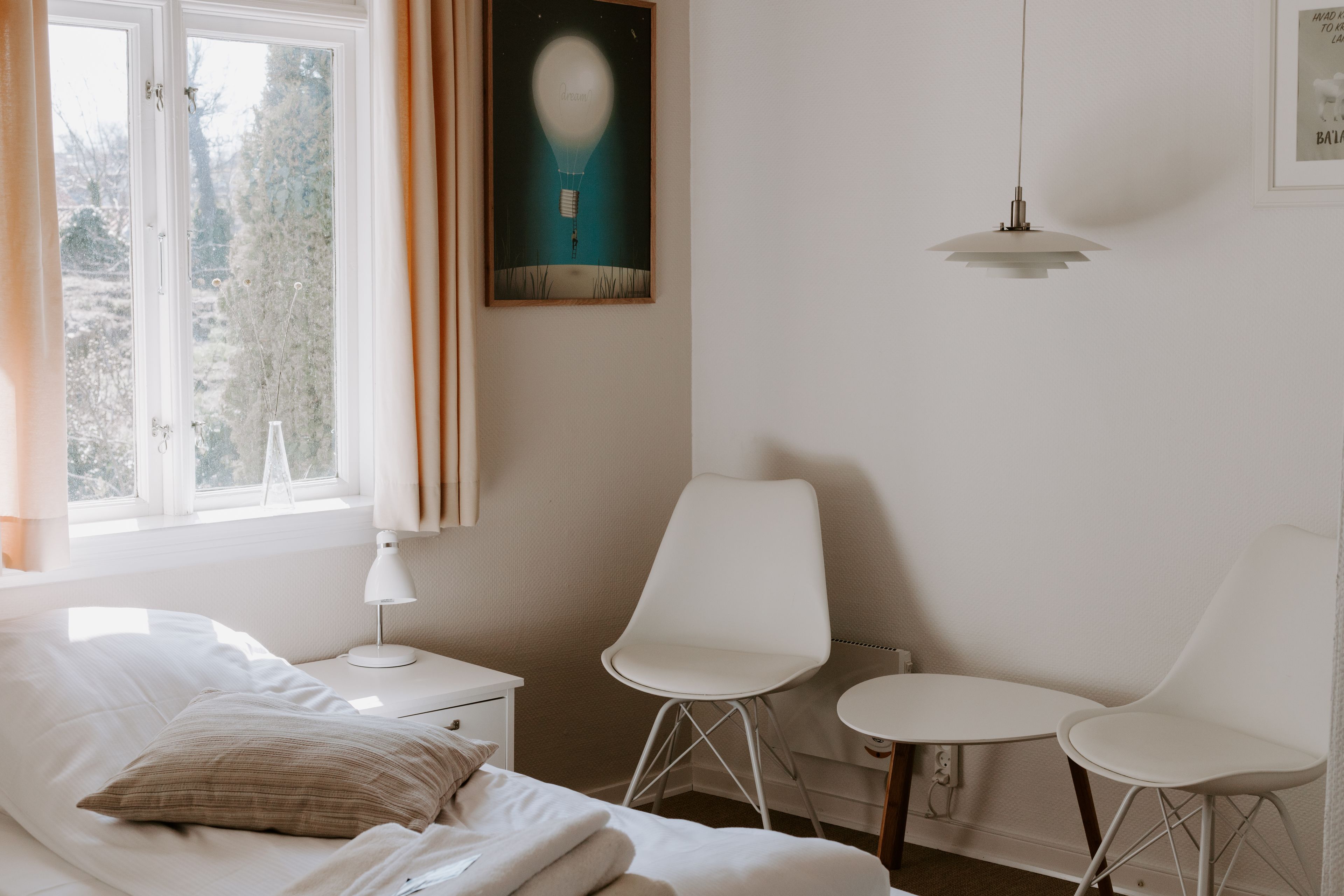 Un dormitorio con cama, mesita de noche, escritorio, dos sillas, lámpara colgante y un cuadro en la pared.