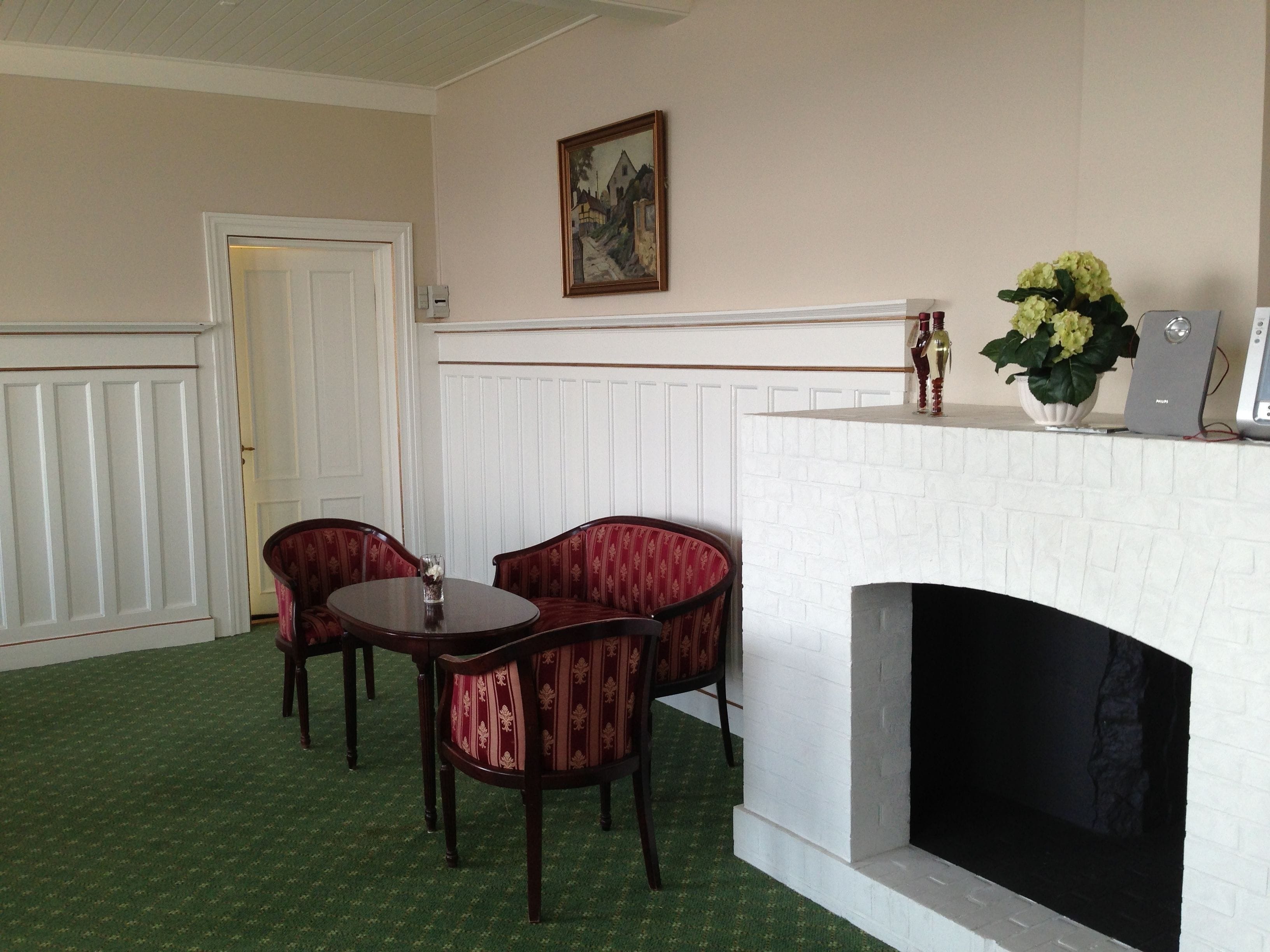 A room with a white brick fireplace, two red upholstered chairs, a small table, and a green carpet.