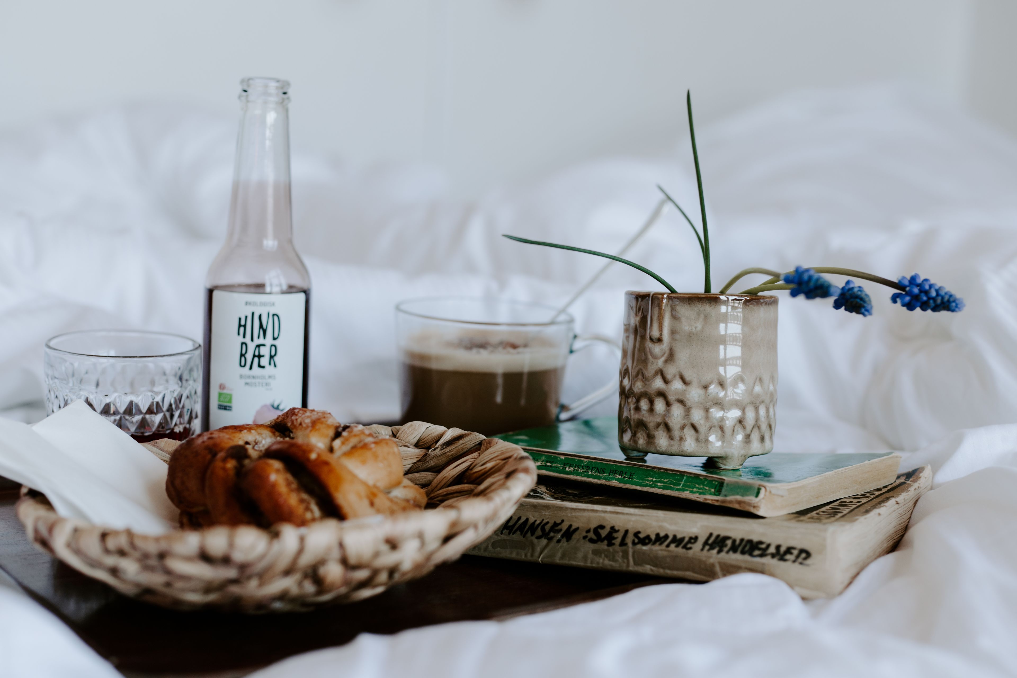 A cozy setting with a bottle labeled "HIND BEER," a glass of beer, a mug, a basket of pastries, and a book on a bed.