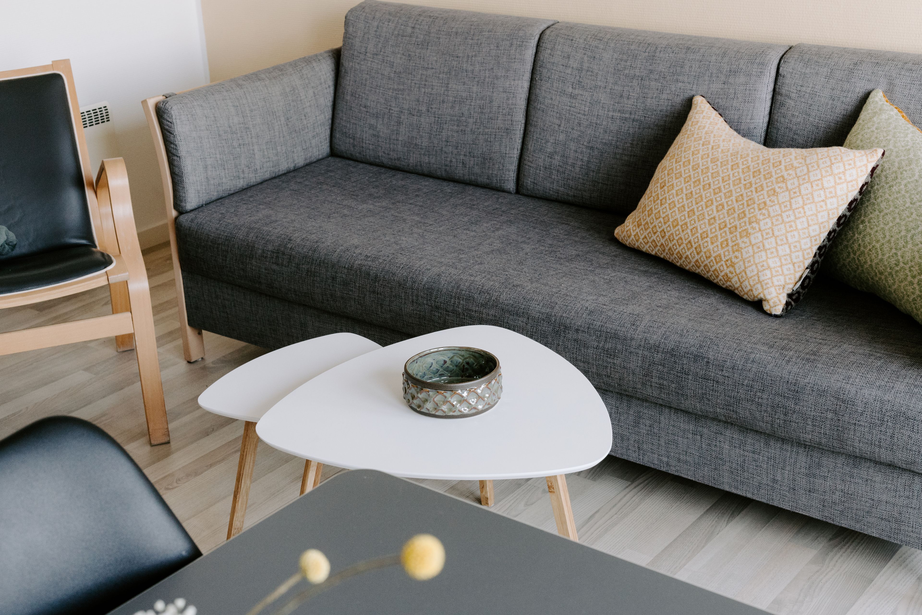 A modern living room with a grey sofa, two white side tables, a black chair, and a decorative bowl on one of the tables.