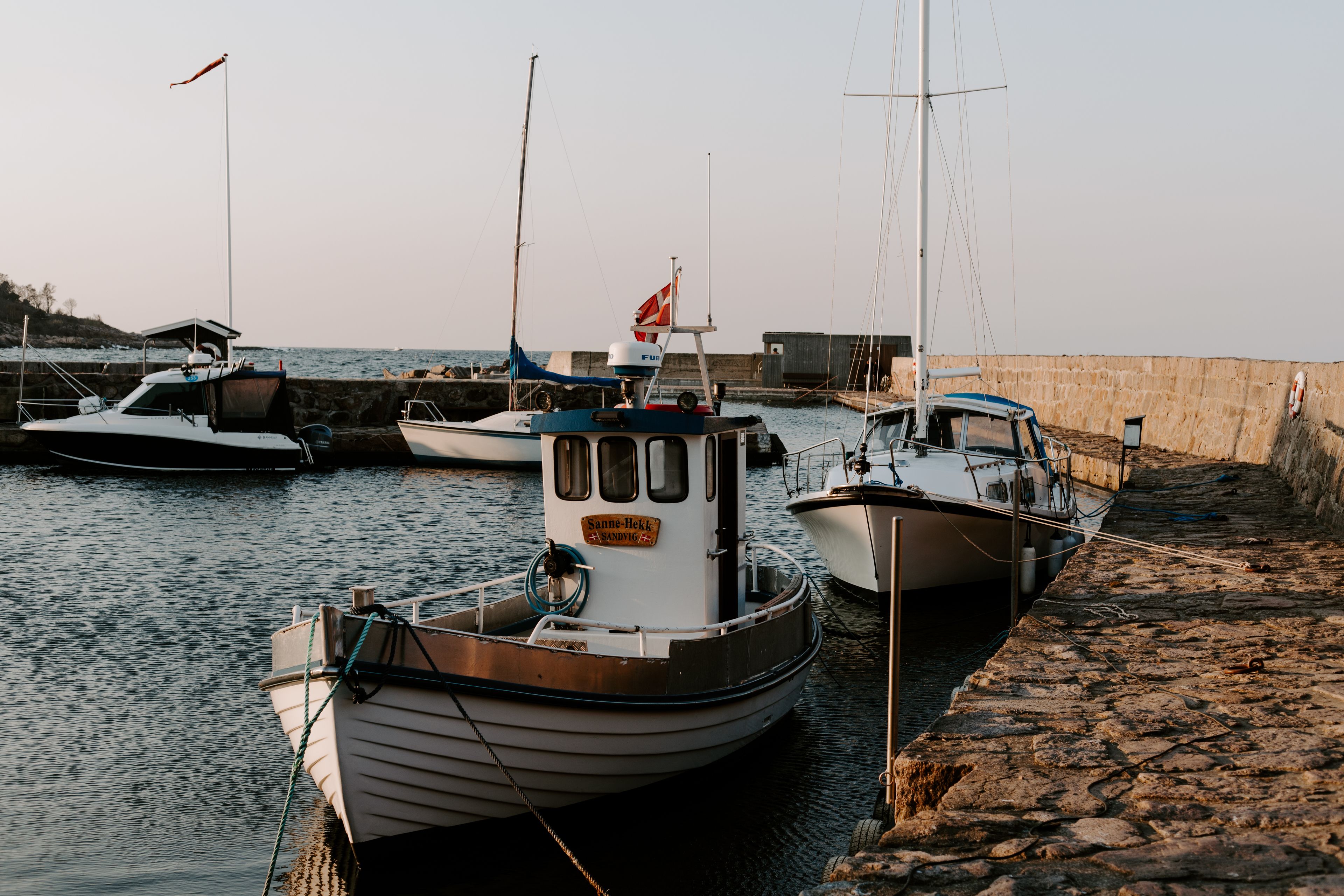 A row of boats docked at a marina with a stone wall and calm water.