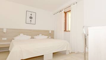 A bedroom with a bed, a nightstand, a wall-mounted light, a window with curtains, and a refrigerator.