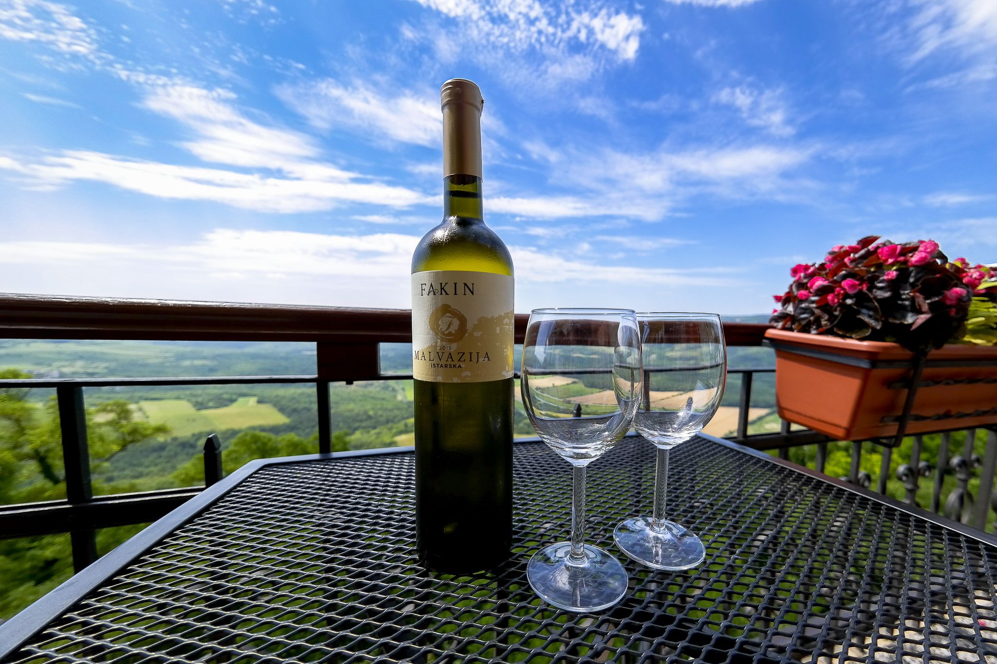 A bottle of Fakin Malvasija wine and two glasses on a metal table with a scenic view.