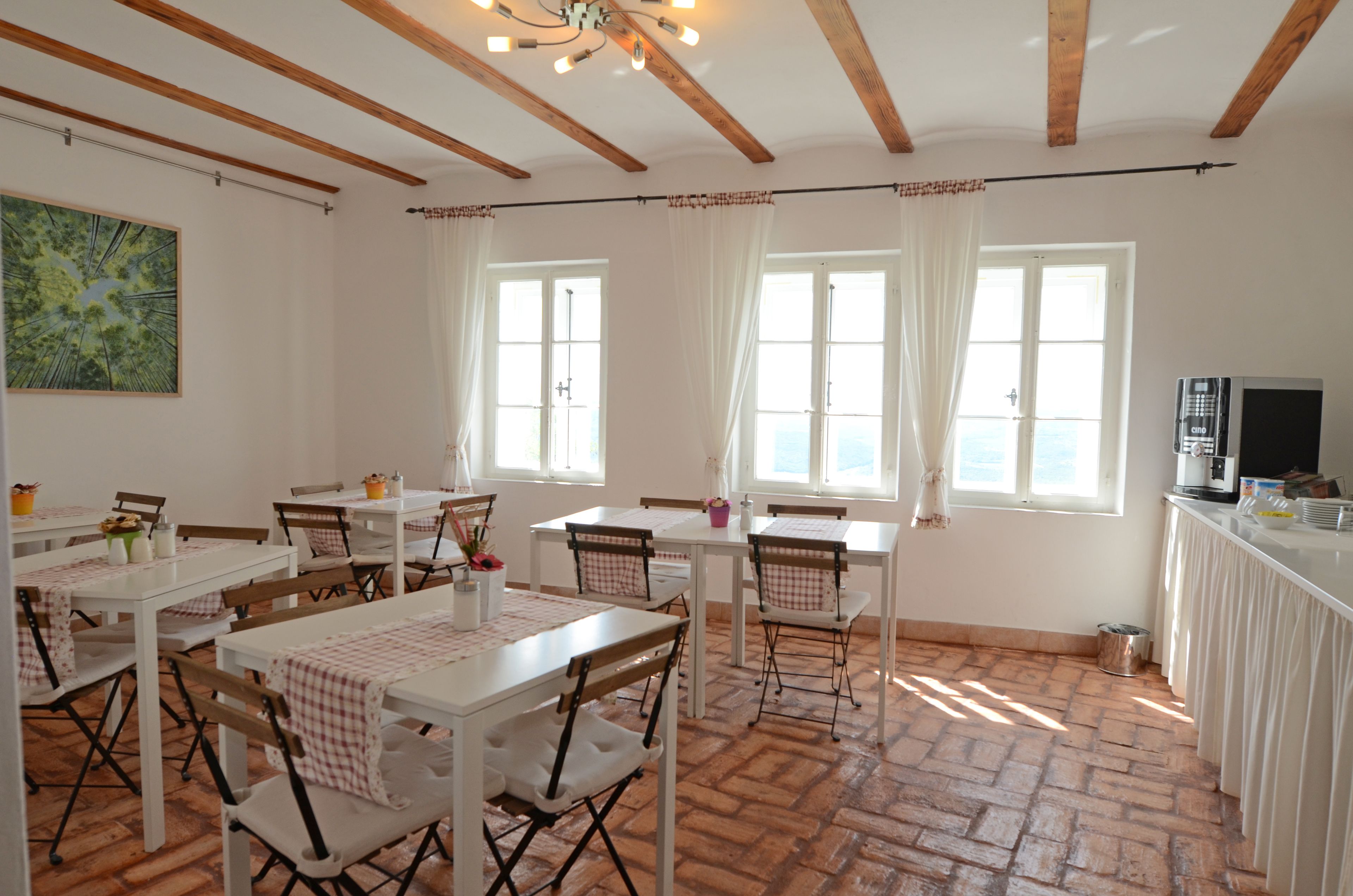 A dining area with white tables and chairs, large windows with curtains, and a kitchen area with a coffee machine.