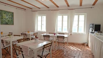 Un comedor con mesas y sillas blancas, ventanas grandes con cortinas, y una zona de cocina con una máquina de café.