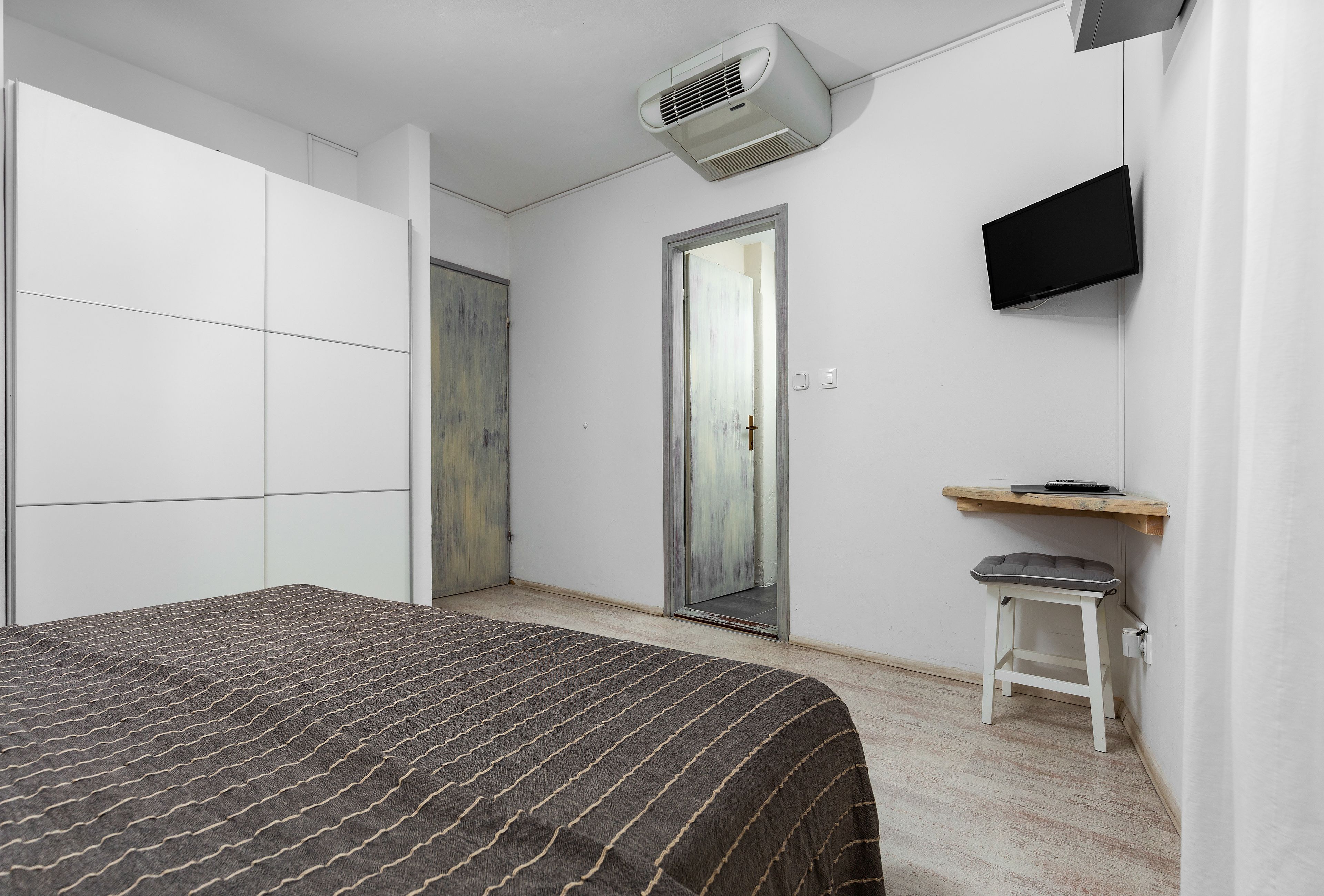 A bedroom with a bed, a TV mounted on the wall, a small stool, and a door leading to another room.