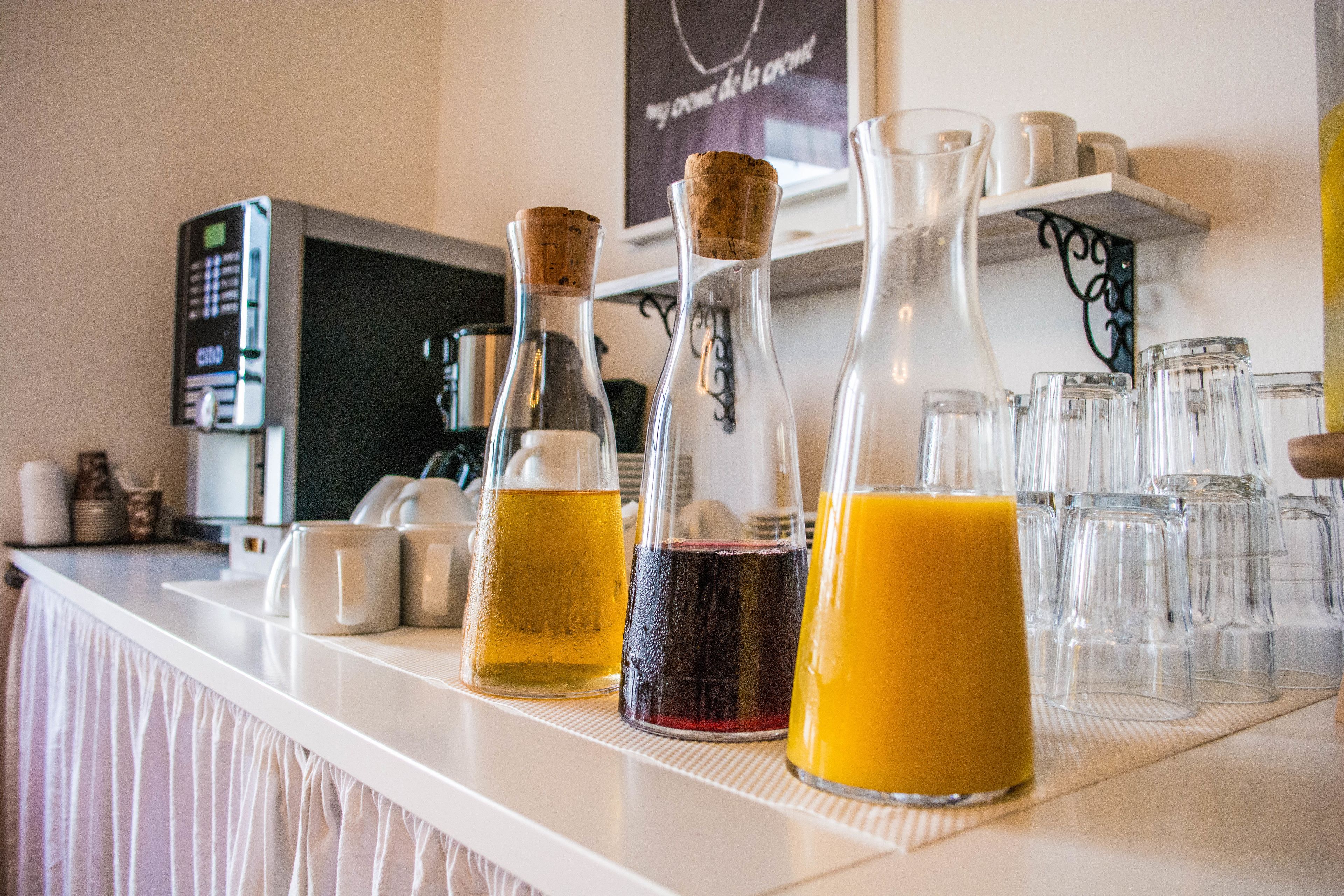 Zona de bar con tres botellas de vidrio llenas de bebidas, una máquina de café y una estantería con tazas y un cartel.