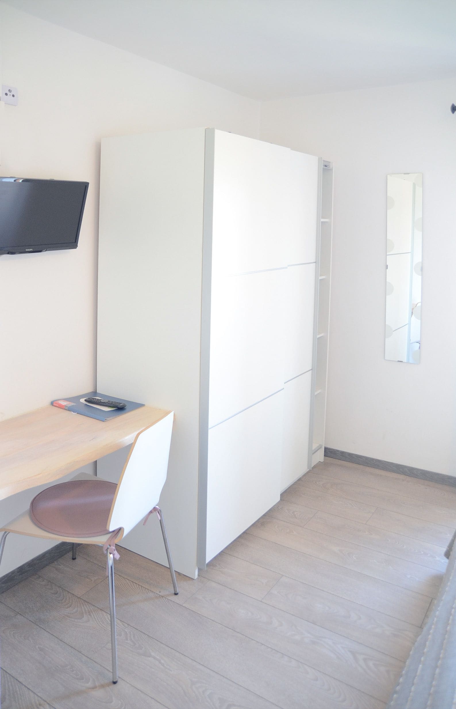 A room with a wooden desk, a chair, a TV mounted on the wall, and a white wardrobe.