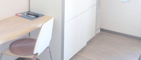 A room with a wooden desk, a chair, a TV mounted on the wall, and a white wardrobe.