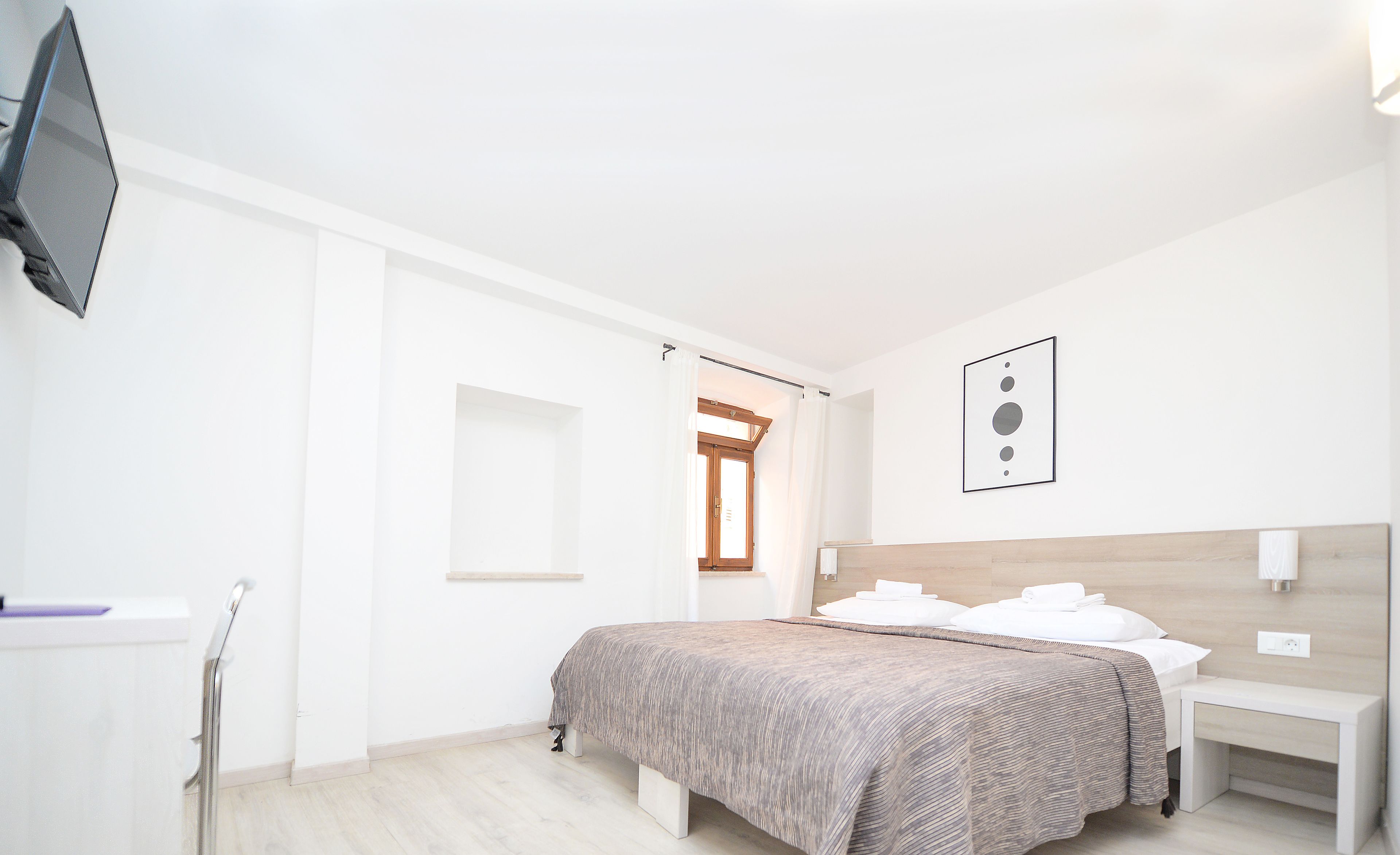 A modern bedroom with a bed, bedside tables, a TV mounted on the wall, and a window with curtains.