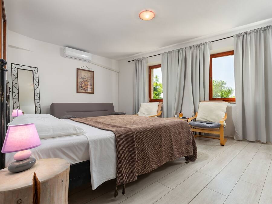Un dormitorio con cama, lámpara de noche, silla y ventana con cortinas.