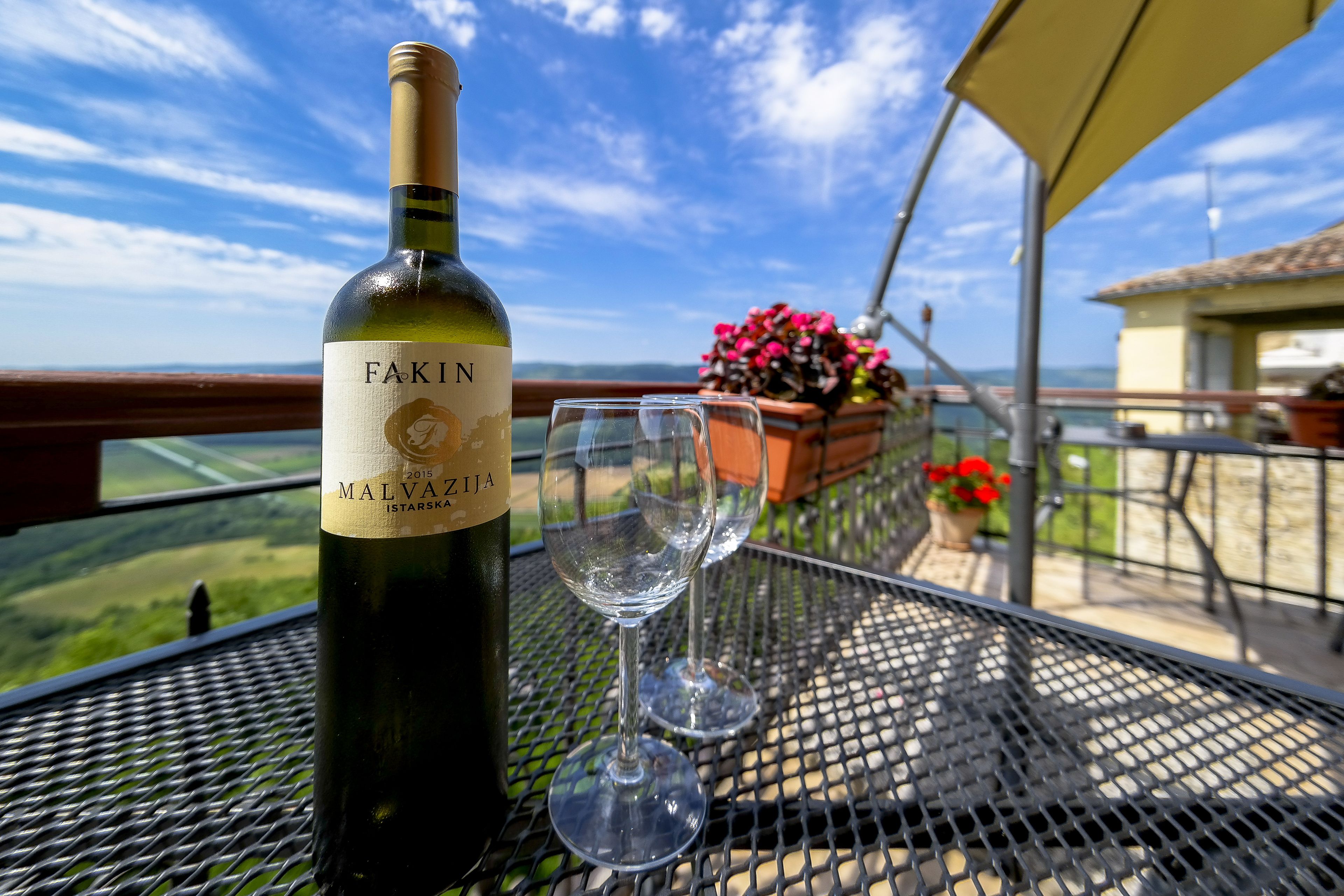 Una botella de vino Malvasia Istarska sobre una mesa metálica con dos copas de vino vacías, en un balcón con vistas al campo.