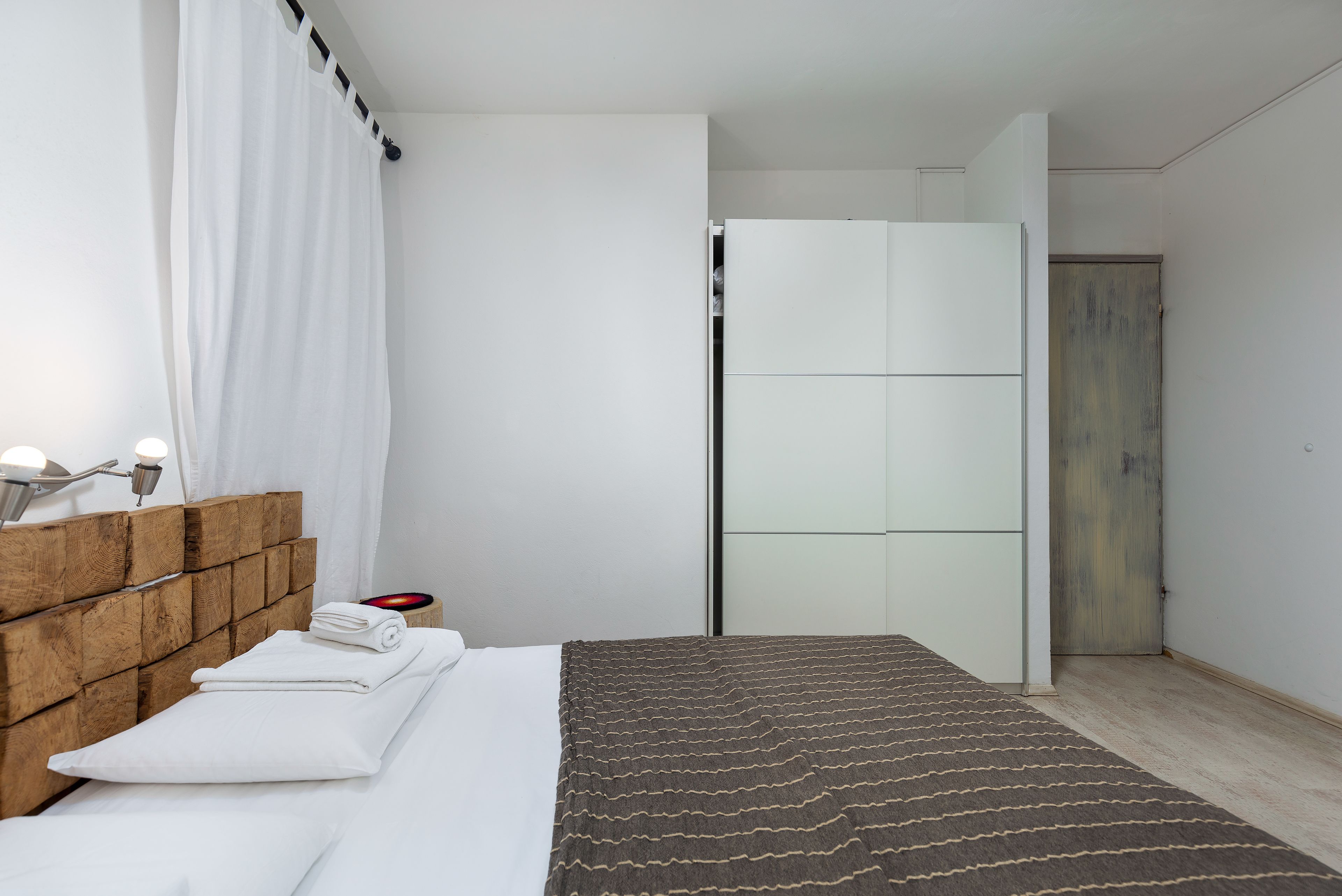 A modern bedroom with a wooden headboard, a neatly made bed, a bedside table with a lamp, and a wardrobe.