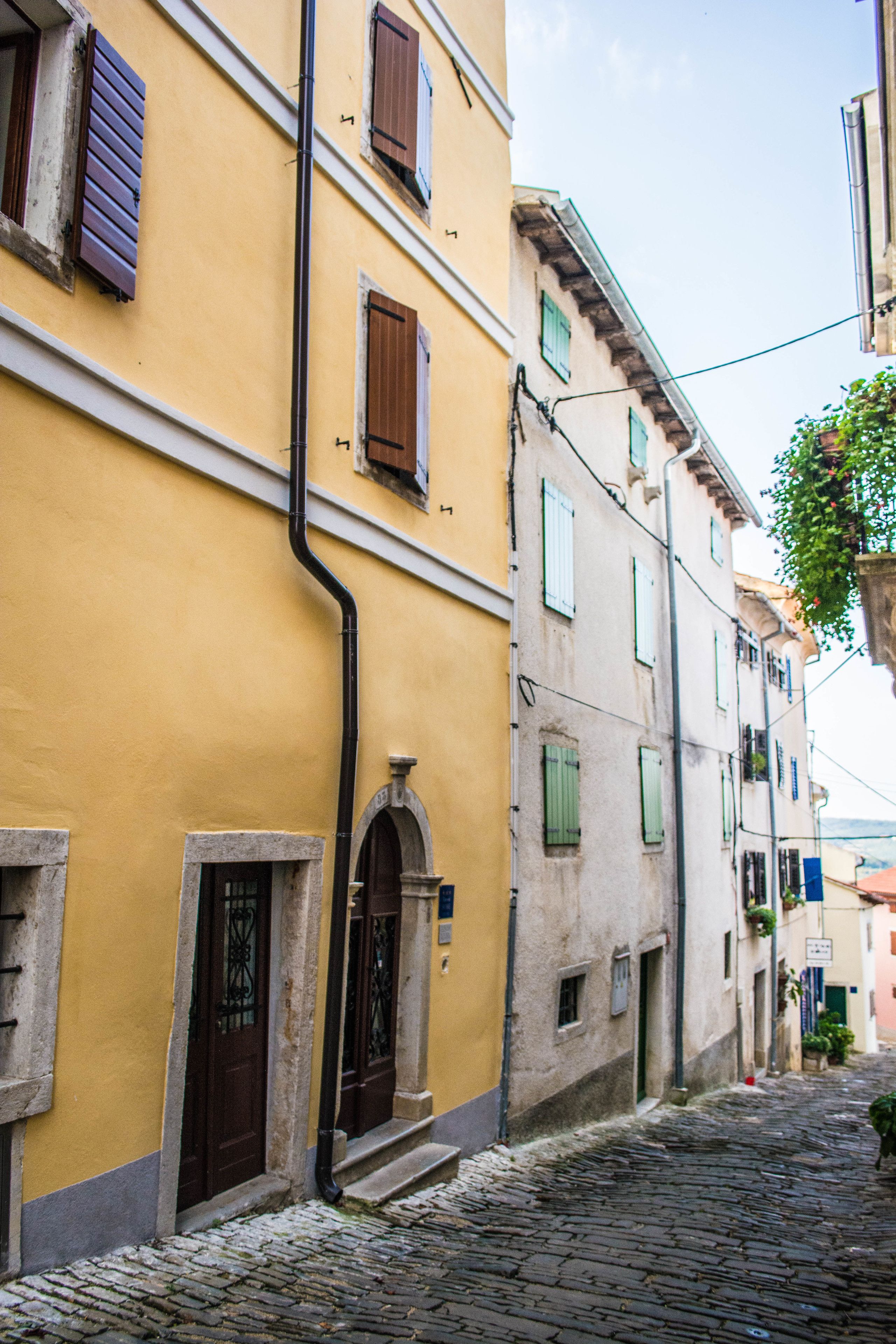 A narrow cobblestone street flanked by yellow buildings with green shutters and a black pipe running along the wall.
