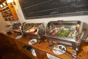A buffet setup with various food items in metal containers, utensils on plates, and a chalkboard in the background. - Hotel Chata Orlik all inclusive (Pec pod Snezkou)