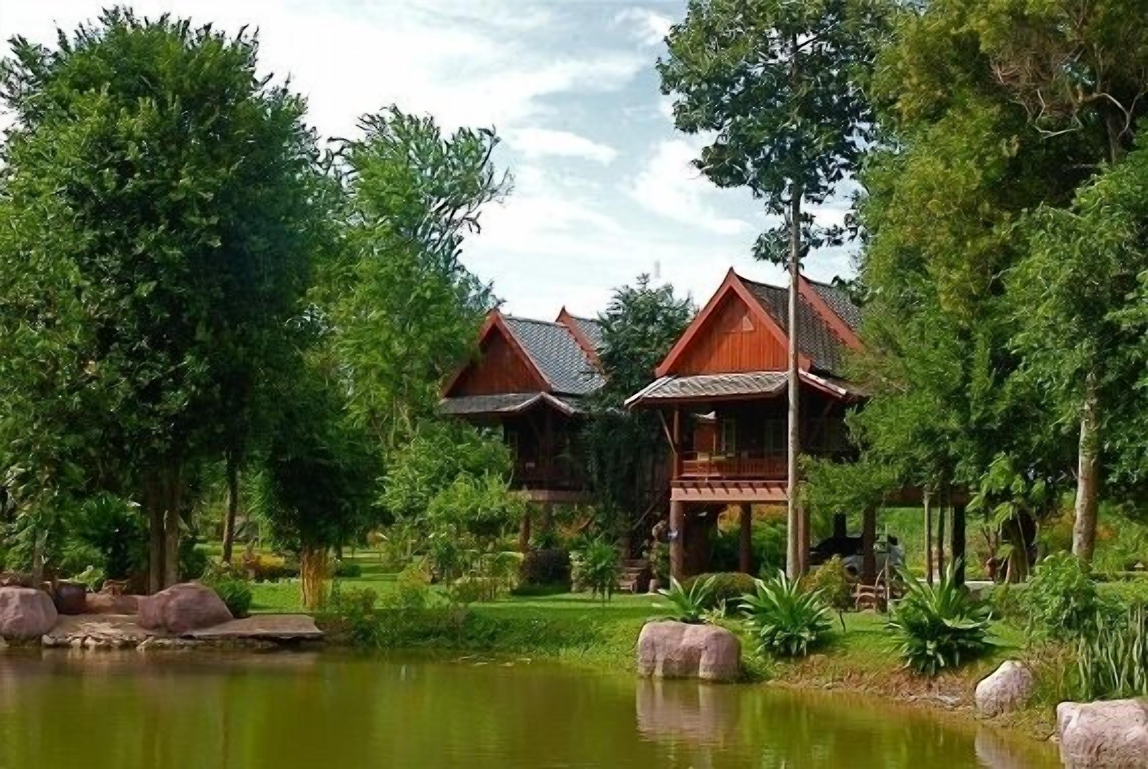 A traditional wooden house with a thatched roof, surrounded by lush greenery and a pond.