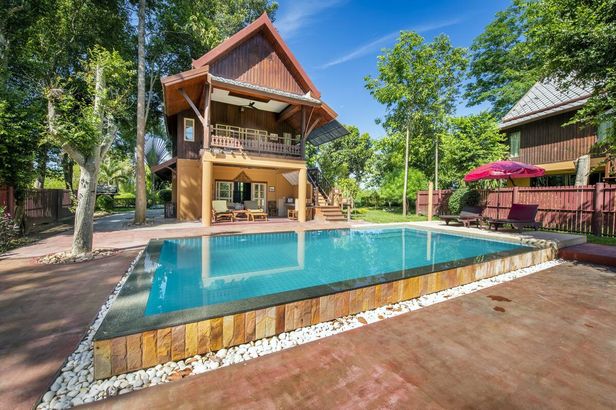 Une maison en bois à deux étages, dotée d’une piscine, de mobilier d’extérieur et d’un parasol rouge.