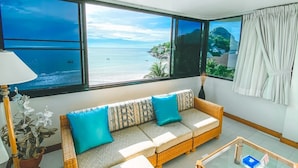 Ein Zimmer mit großem Fenster mit Blick auf einen Strand, einer Couch mit gemusterten Kissen, einer Lampe und einer Topfpflanze.