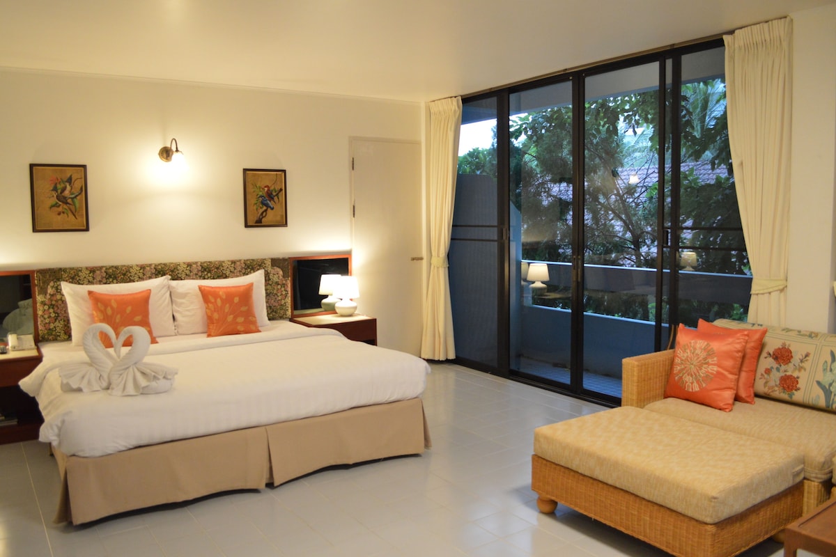 A hotel room with a large bed, a sofa, a nightstand, a lamp, and a window with a view of trees.