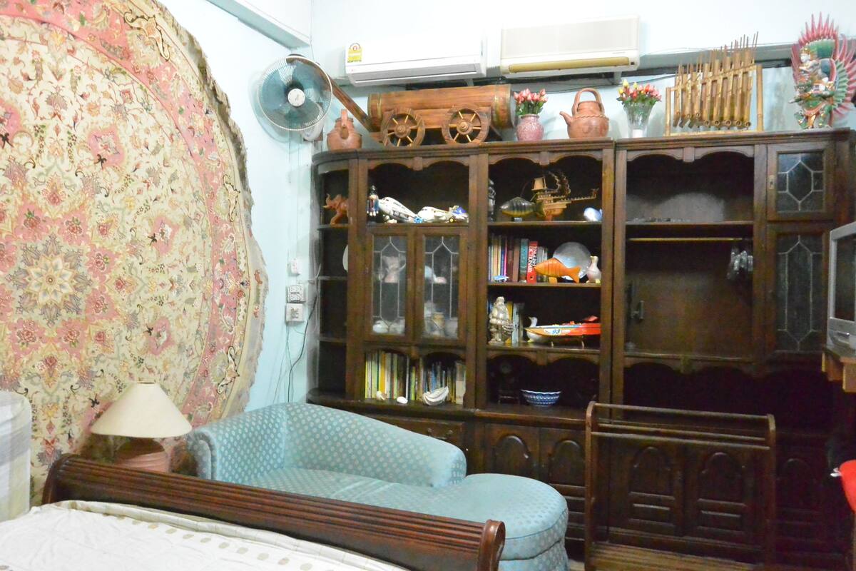 Ein Zimmer mit einem gemusterten Vorhang, einem Holzschrank mit diversen Gegenständen, einem blauen, gemusterten Sofa und einem hölzernen Bett.