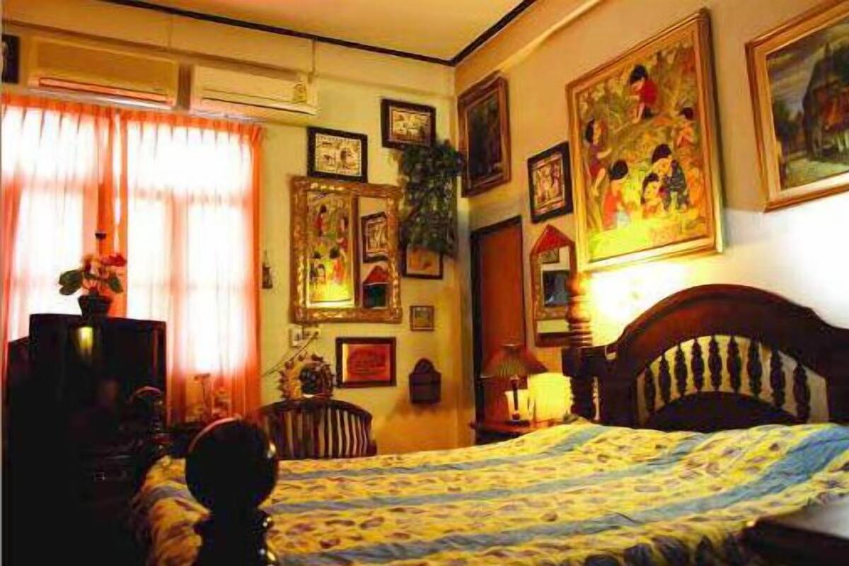 Ein Schlafzimmer mit einem Bett, einem Nachttisch, einer Lampe und verschiedenen Wanddekorationen, darunter gerahmte Bilder und eine Pflanze.