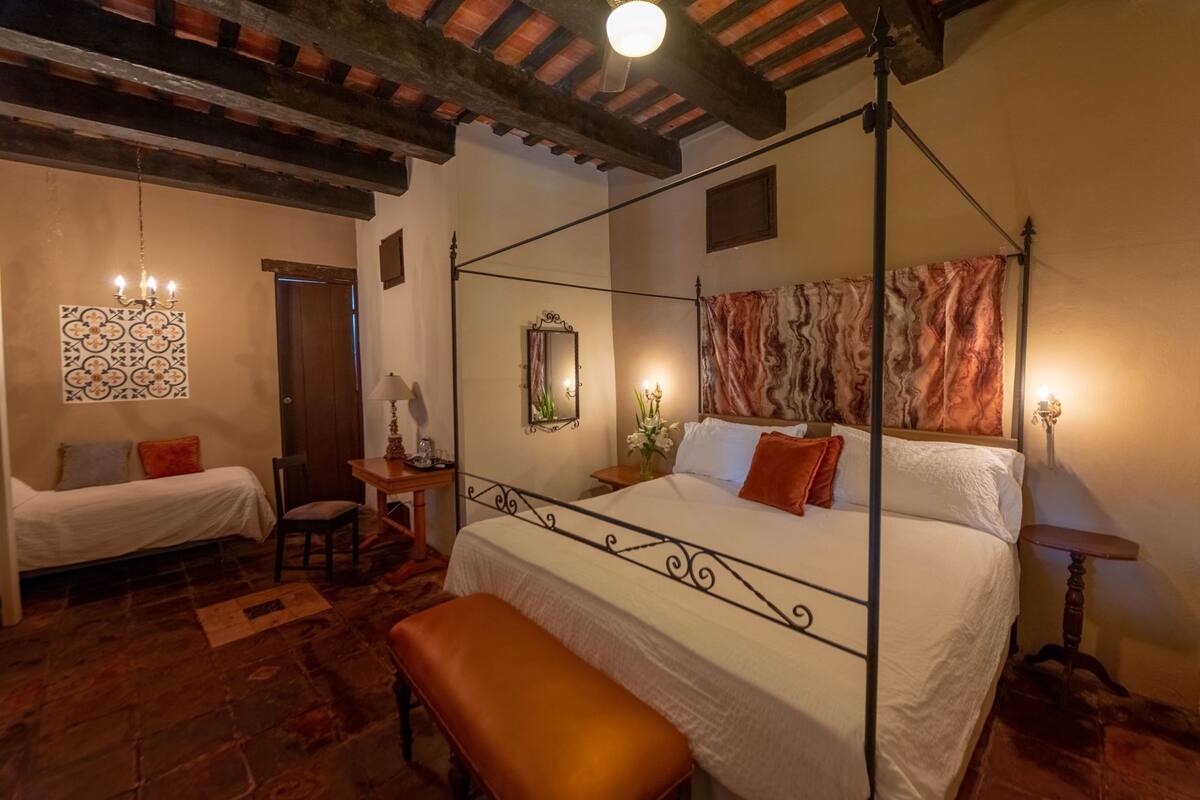 A bedroom with a four-poster bed, a bench, a desk with a lamp, a mirror, and a patterned wall decoration.