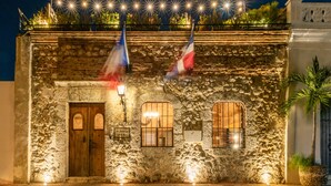 A stone building with a wooden door, two windows with shutters, and flags. - El Beaterio (Santo Domingo)
