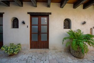 Una entrada de edificio con puerta de madera, dos ventanas arqueadas con celosías metálicas y plantas en macetas en el suelo.