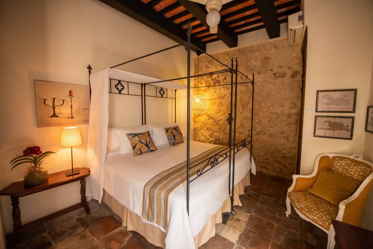 A four-poster bed with a canopy, a bedside table with a lamp, a chair, and a stone wall.