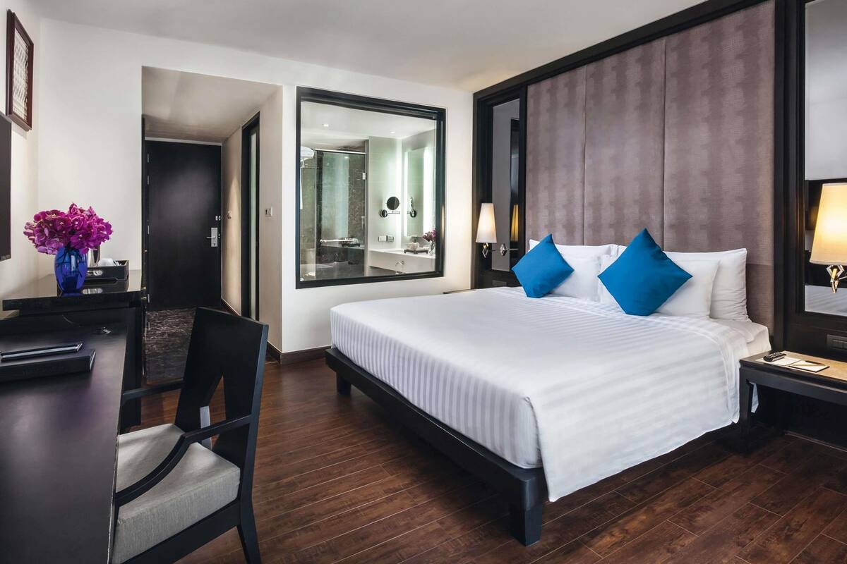 A hotel room with a large bed, a desk, a chair, a mirror, and a bathroom visible through the glass door.